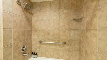 Baño con azulejos color beige, una bañera, un cabezal de ducha manual, una barra de apoyo y un dispensador de jabón.