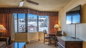 Hotelkamer met een groot raam met uitzicht op een besneeuwde bergstad. Er is een flatscreen televisie aan de muur, een houten bureau met een stoel, en een bank.