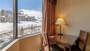 Een hotelkamer met een groot raam dat uitzicht biedt op een besneeuwde berg en gebouwen. Er staat een tafel met een lamp en twee stoelen.