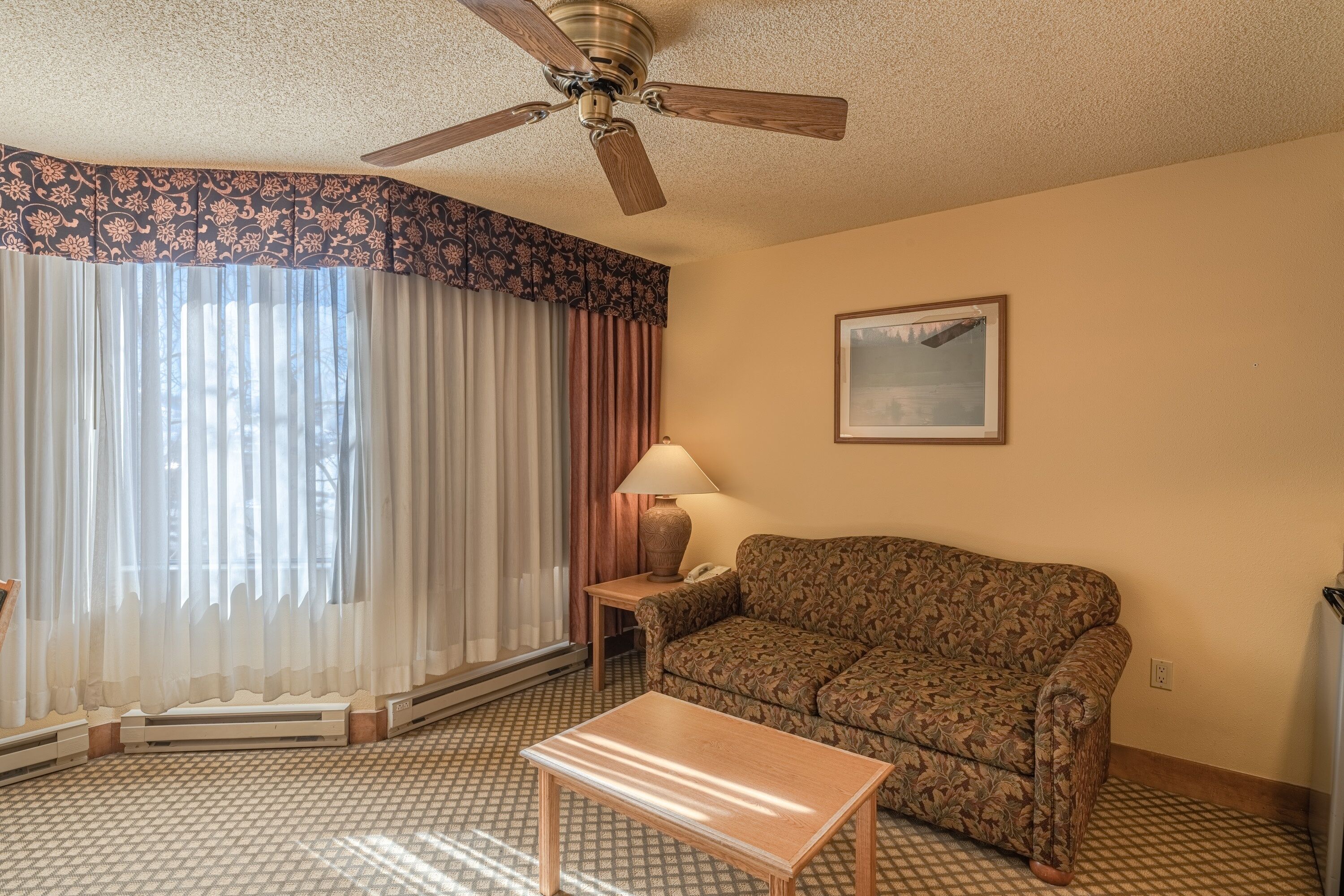 A hotel room with a patterned sofa, a wooden coffee table, a standing lamp, a ceiling fan, and a large window with sheer curtains.