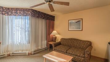 A hotel room with a patterned sofa, a wooden coffee table, a standing lamp, a ceiling fan, and a large window with sheer curtains.