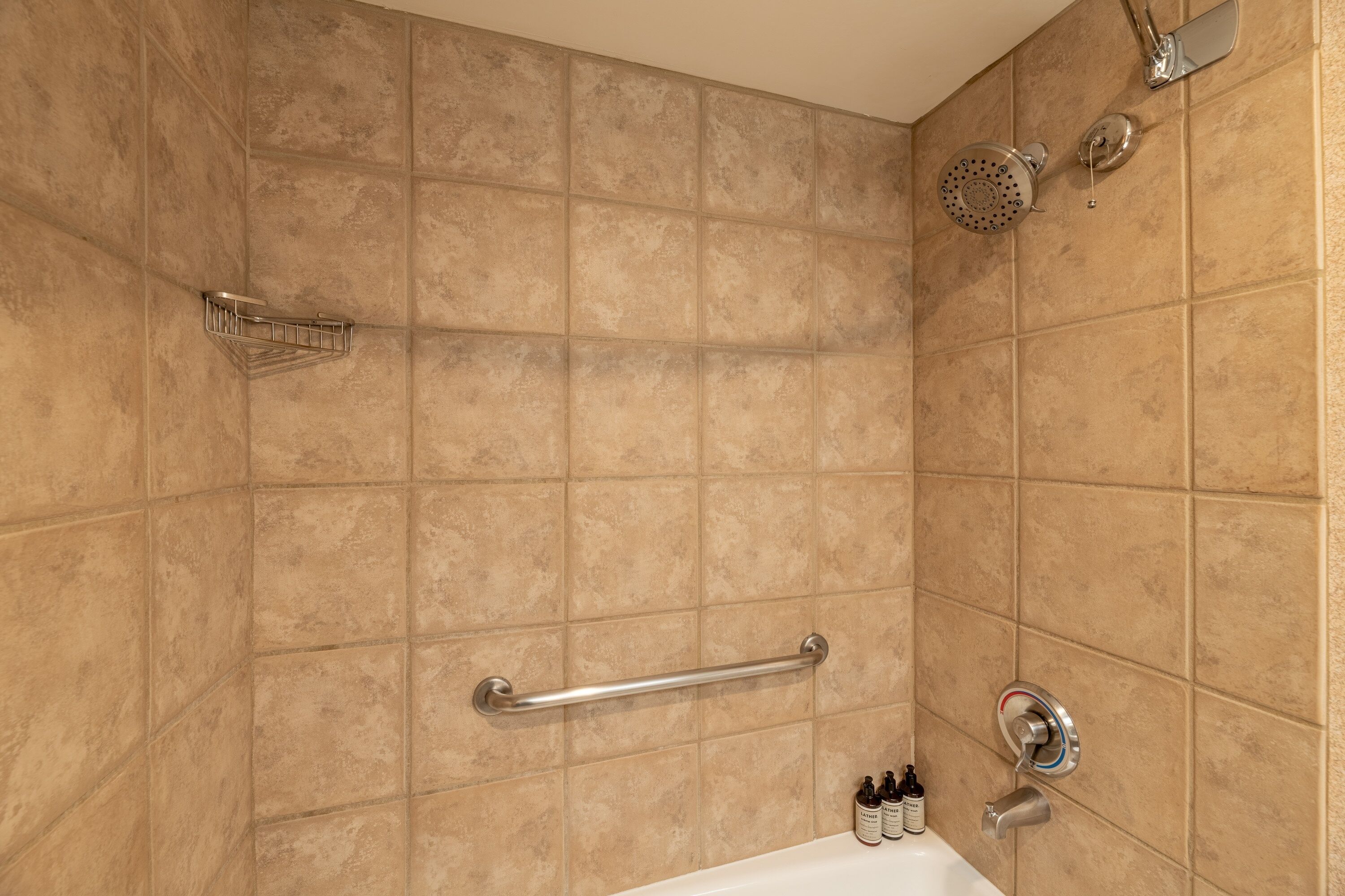 Une salle de bain avec des carreaux de couleur beige, une barre d’appui, une pomme de douche et une bouteille de shampoing.