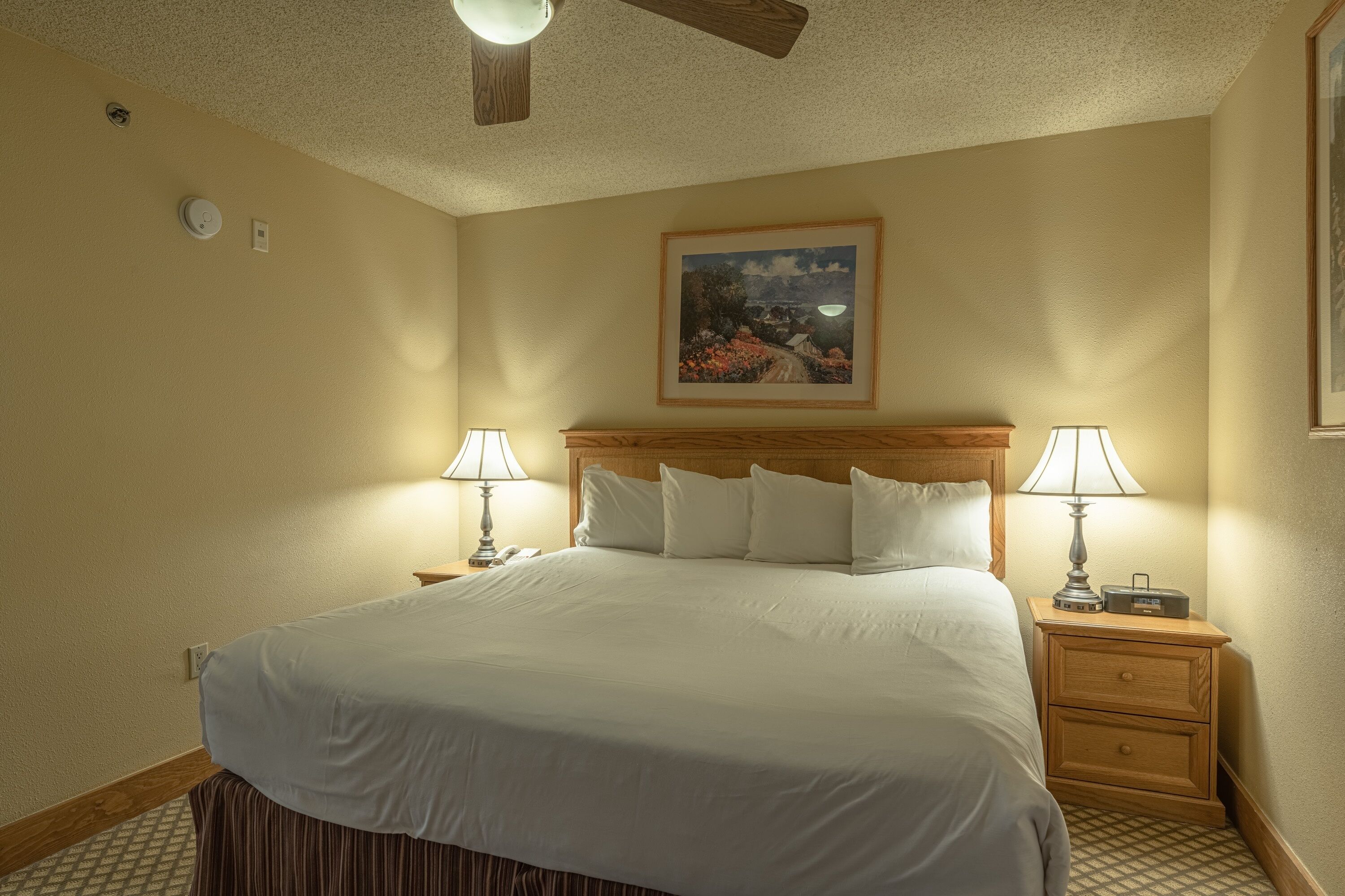 Una habitación de hotel con una cama grande, dos mesitas de noche con lámparas, un ventilador de techo y un cuadro en la pared.
