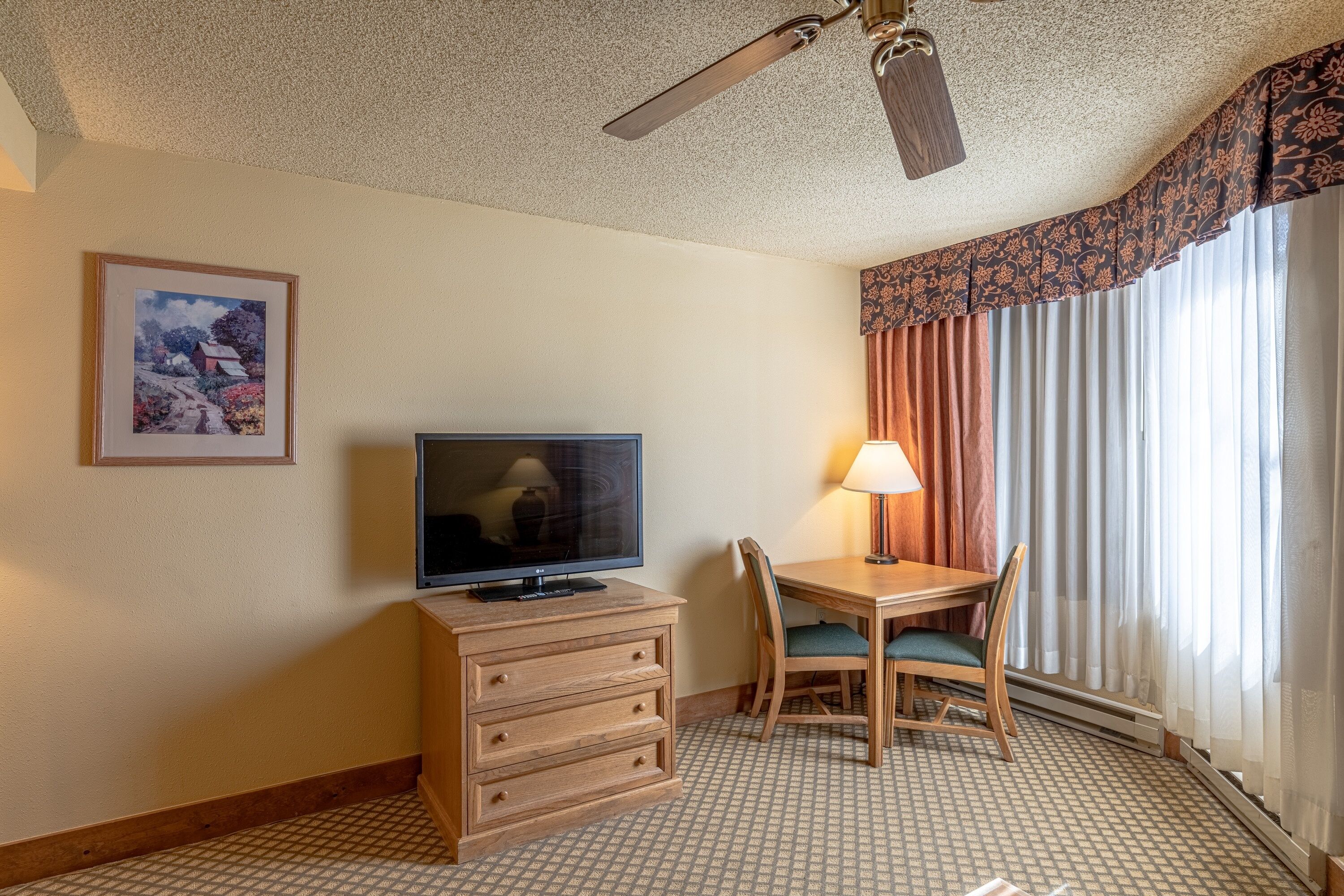 Une chambre d’hôtel avec un téléviseur posé sur une commode en bois, une table à manger avec des chaises, un ventilateur de plafond et une grande fenêtre avec des rideaux.