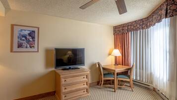 Une chambre d’hôtel avec un téléviseur posé sur une commode en bois, une table à manger avec des chaises, un ventilateur de plafond et une grande fenêtre avec des rideaux.