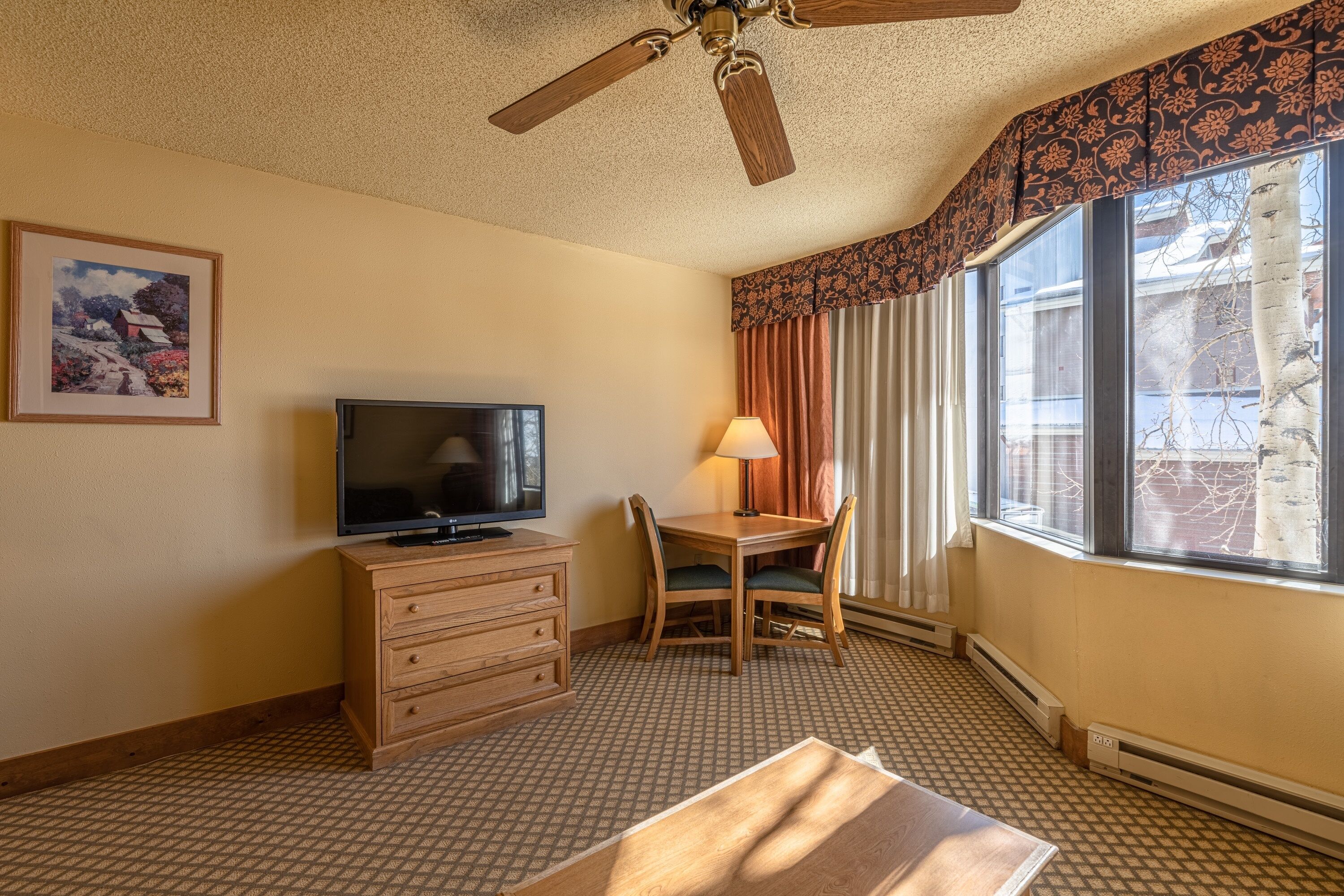 A hotel room with a ceiling fan, a television on a wooden dresser, a dining table with chairs, a window with curtains, and a framed picture on the wall.