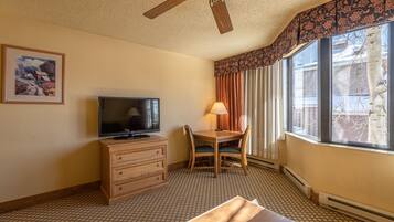 A hotel room with a ceiling fan, a television on a wooden dresser, a dining table with chairs, a window with curtains, and a framed picture on the wall.