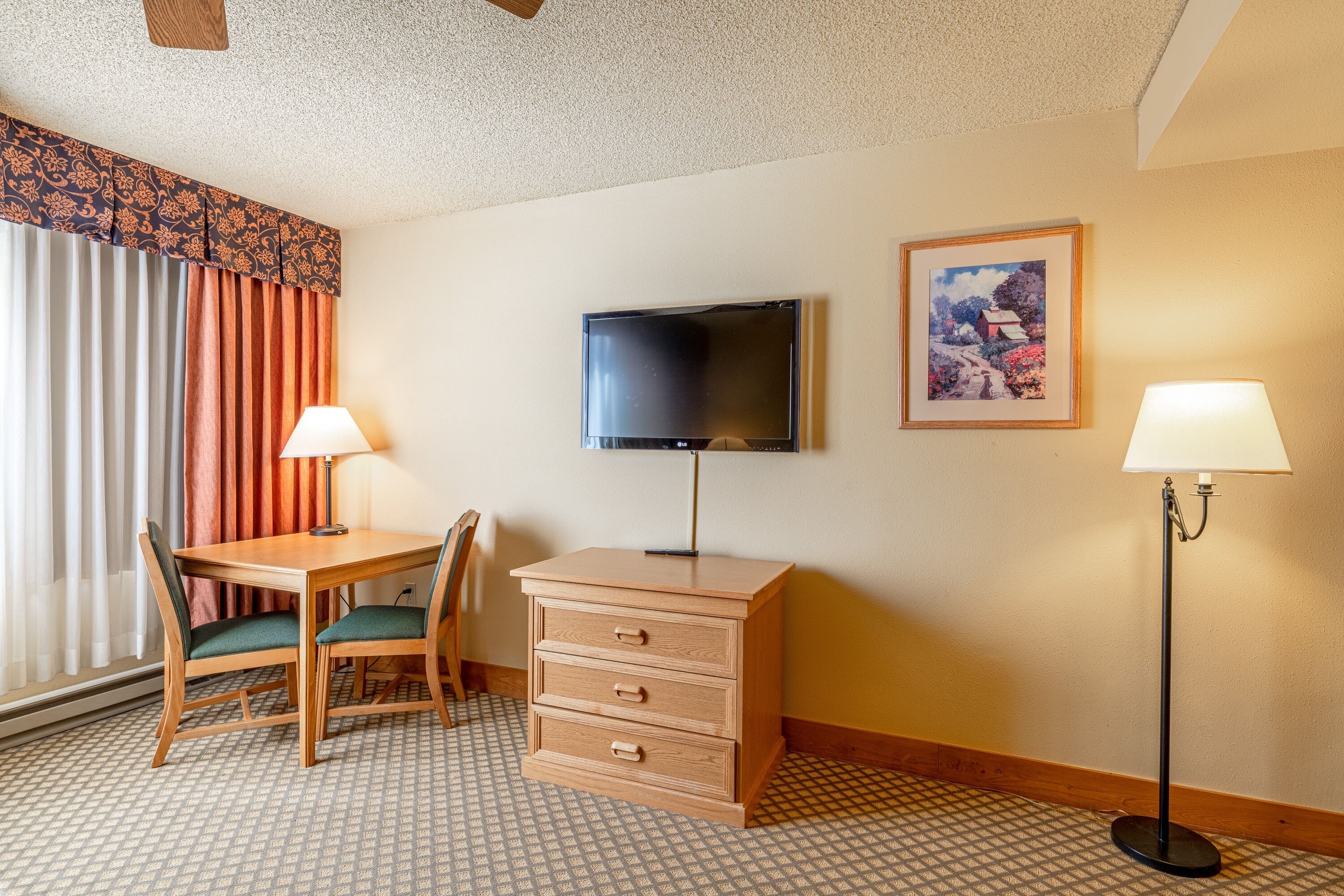 A hotel room with a wooden desk, two chairs, a television, a dresser, two lamps, and a patterned floor.