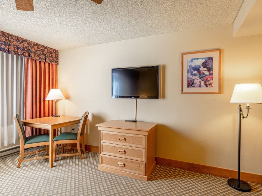 A hotel room with a wooden desk, two chairs, a television, a dresser, two lamps, and a patterned floor.