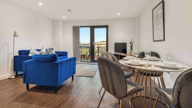 A modern living room with a blue sofa, a dining table set for four, and a large window with a view of buildings. - Stay Zeni, Ouse Court York Stylish 2 Bed Apartment (York)