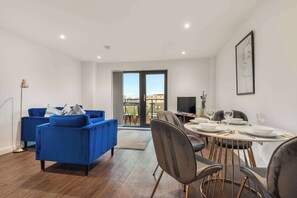 A modern living room with a blue sofa, a dining table set for four, and a large window with a view of buildings. - Stay Zeni, Ouse Court York Stylish 2 Bed Apartment (York)