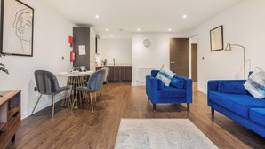 A modern living room with a blue sofa, a dining area with a table and chairs, and a kitchen in the background. - Stay Zeni, Ouse Court York Stylish 2 Bed Apartment (York)