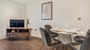 A modern dining area with a round table set for six, surrounded by dark chairs with gold legs. A flat-screen TV is mounted on a wooden console unit with drawers. - Stay Zeni, Ouse Court York Stylish 2 Bed Apartment (York)