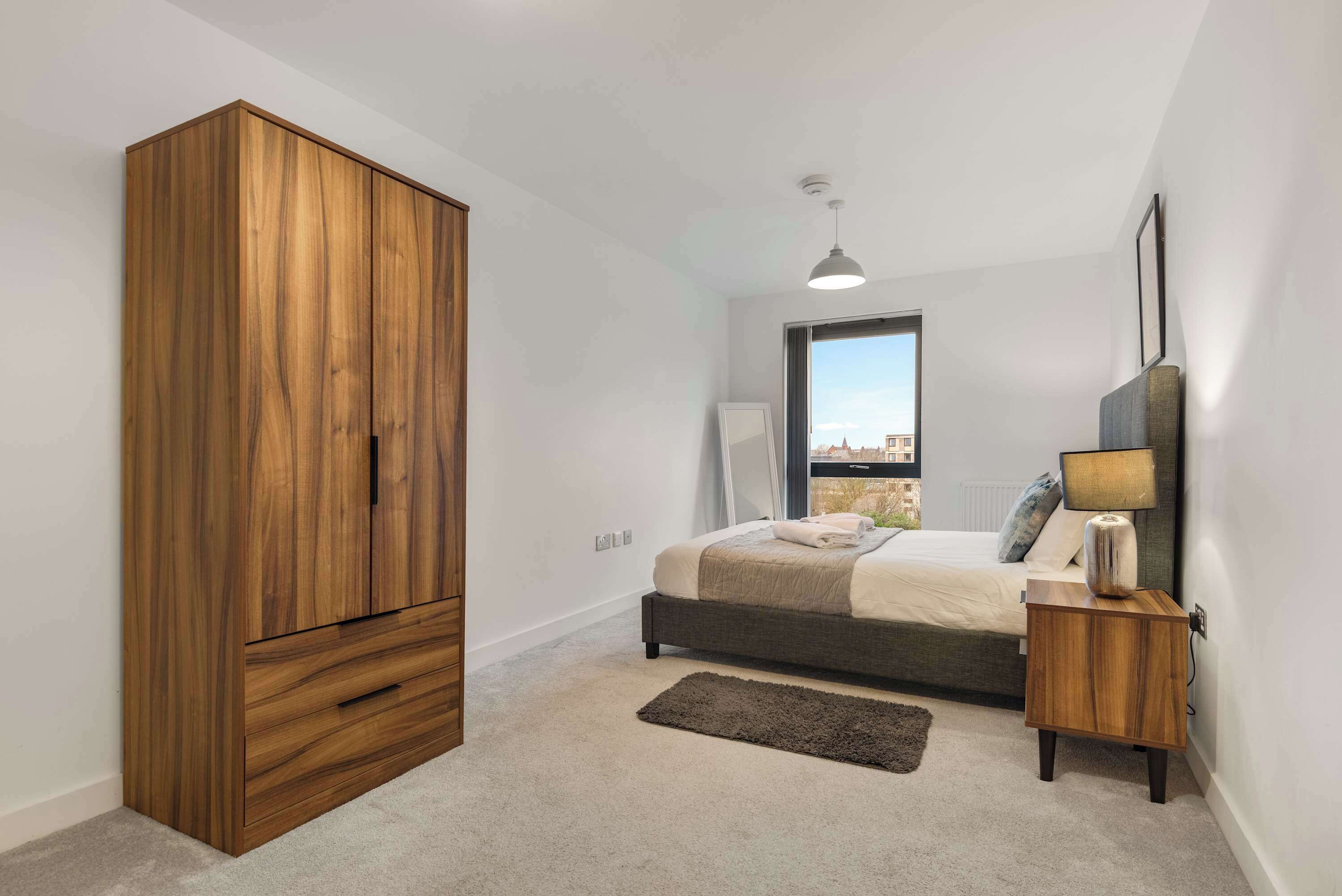 A bedroom with a large wooden wardrobe, a bed with a grey headboard, a bedside table with a lamp, and a view of the city through a window.