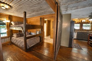 A cabin interior with a bunk bed, wooden walls, a kitchen area, and a view through a window.