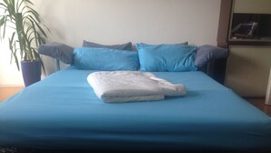 A bed with a blue mattress and pillows, a folded white blanket, and a potted plant to the left.