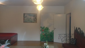 A spacious room with a large window, a painting on the wall, a door with a green floral design, a chair, and a small table with a plant.