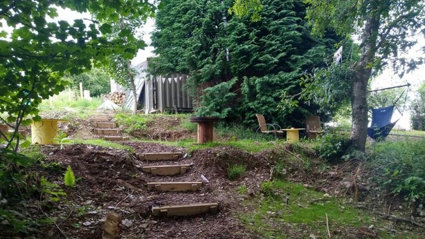 A garden with a wooden staircase leading up a slope, surrounded by greenery and a wooden fence. - Pilgrims Shepherd's Hut - Goosewell Glynn Glamping (Callington)