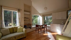 A spacious living area with a sofa, a dining table, and chairs, a kitchen with white cabinets, and large windows offering views of greenery. - A delightful studio in idyllic Mapua Village, perfect from which to explore the Tasman region (Mapua)