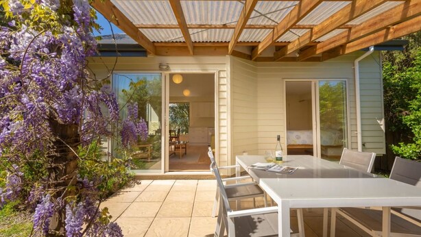 A modern outdoor dining area with a glass table and chairs, a pergola overhead, and purple wisteria flowers. - A delightful studio in idyllic Mapua Village, perfect from which to explore the Tasman region (Mapua)