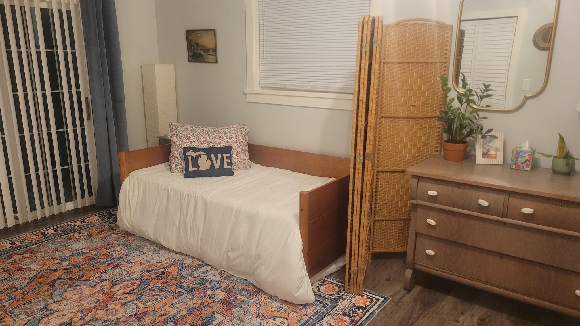 A bedroom with a bed, a dresser, a mirror, a window with blinds, a door with a curtain, and a patterned rug.