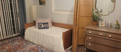 A bedroom with a bed, a dresser, a mirror, a window with blinds, a door with a curtain, and a patterned rug.