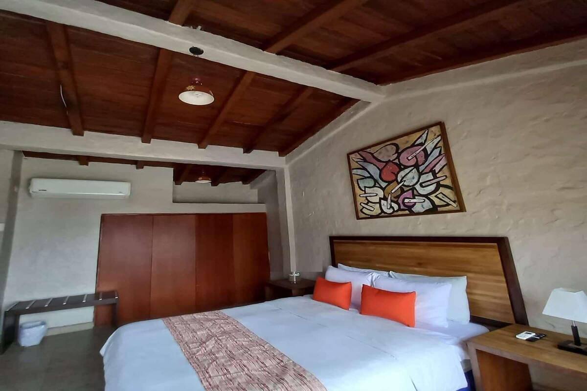 Ein Hotelzimmer mit Holzboden, einem Bett mit weißer Bettwäsche und orangefarbenen Kissen, einem Nachttisch mit Lampe und einem Wandbild.