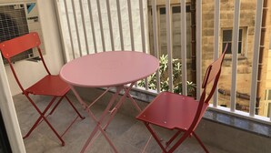A balcony with a round table and two red chairs, a white railing, and a view of a building with windows and a door. - Sliema Beach Apartment (Sliema)
