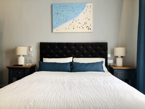 Cama bem arrumada com roupa de cama branca e azul, ladeada por duas mesas de cabeceira com abajures, uma obra de arte na parede retratando uma cena de praia, e uma parede lisa.