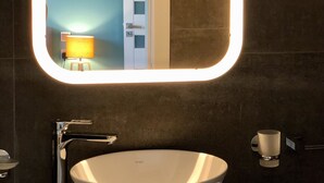A bathroom with a white sink, a mirror with a light, and a door in the background. - Sliema Beach Apartment (Sliema)