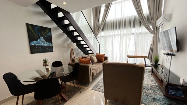 Un salon moderne avec une table à manger en verre, des chaises noires, un canapé et un fauteuil marron. Un escalier mène à un autre niveau.