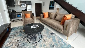 Un salon moderne avec un canapé, des fauteuils et une table basse. On aperçoit à l’arrière-plan un coin cuisine avec des placards et des appareils électroménagers.