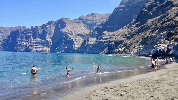 A beach with clear blue water, rocky cliffs, and a boat in the distance. - Luxury and functional sea view apartment (Los Gigantes)