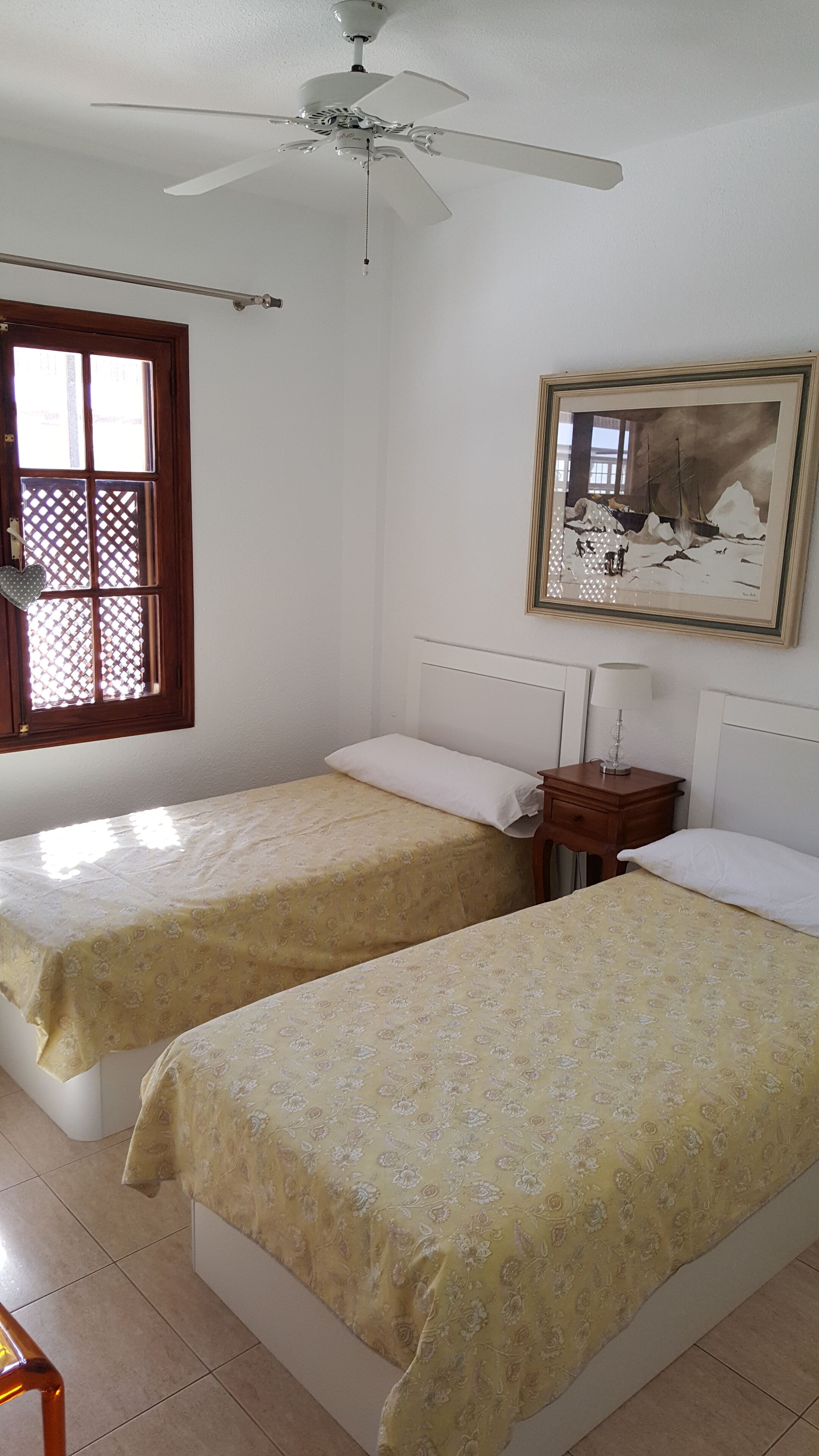 A room with two beds, a ceiling fan, a window with a lattice, and a framed painting on the wall.