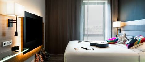 A modern hotel room with a large flat-screen TV, a bed with colorful pillows, a bedside table with a lamp, and a window with curtains.