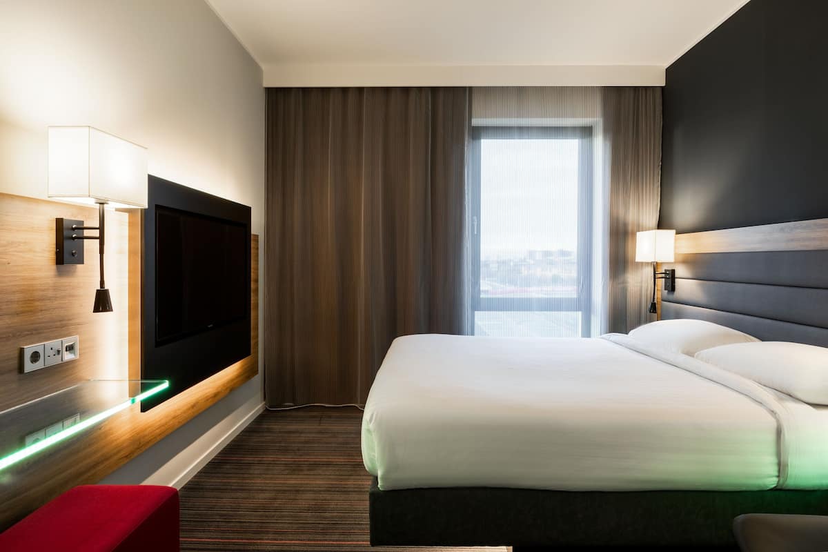 A hotel room with a bed, a TV, a desk with a lamp, and a window with curtains.