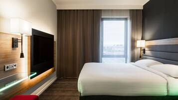 A hotel room with a bed, a TV, a desk with a lamp, and a window with curtains.