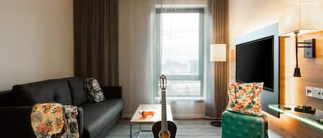 Ein Hotelzimmer mit einer schwarzen Couch, einem kleinen Tisch, einer Gitarre, einem grünen Hocker, einem Fernseher und einem Fenster mit Vorhängen.