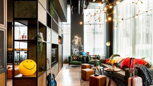 A modern living room with a sofa, ottomans, a coffee table, and a guitar. There is a large window with curtains and a wall-mounted light fixture.