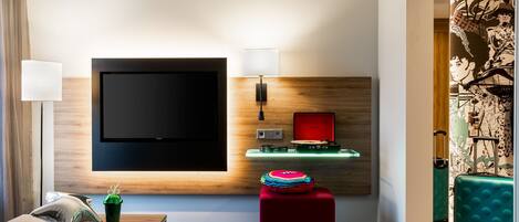 A modern hotel room with a large flat-screen TV mounted on the wall, a wooden accent wall, a small table with a lamp, a red ottoman, and a teal suitcase.
