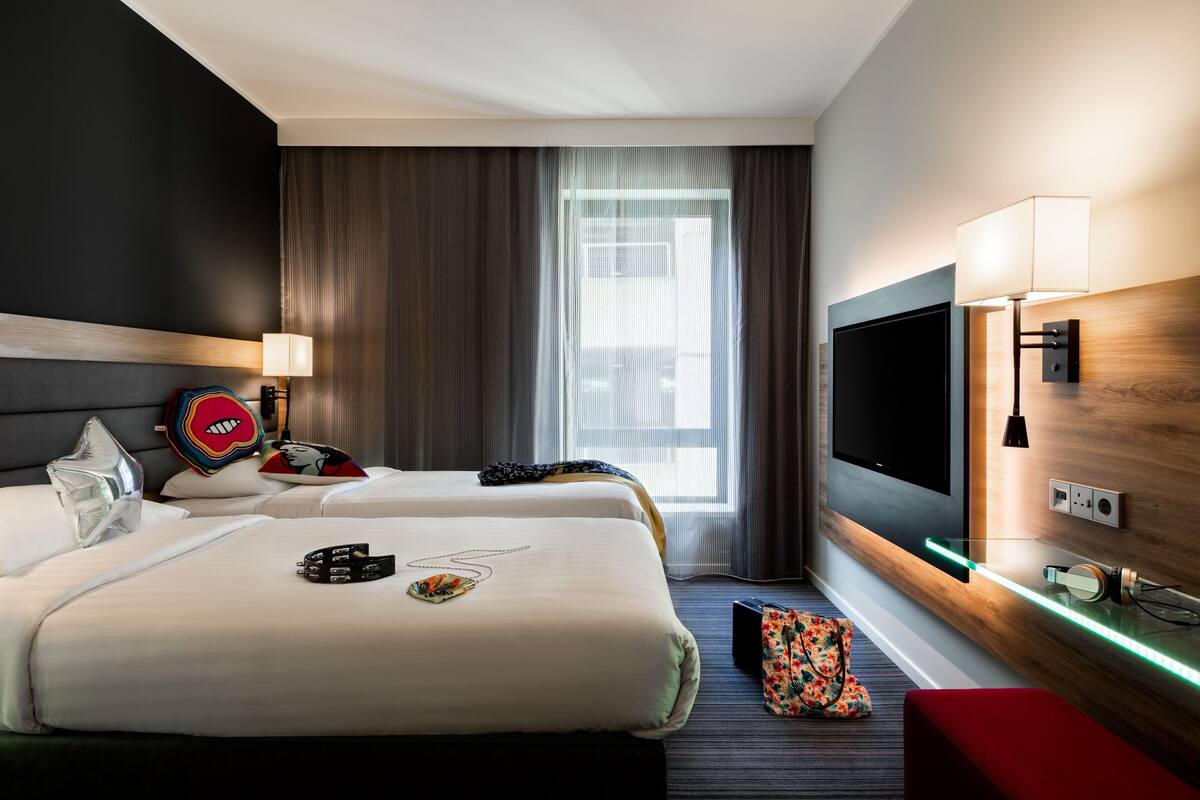 A hotel room with two beds, a TV, a desk, and a window with curtains.
