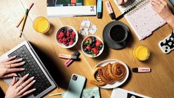A wooden table with a laptop, a smartphone, bowls of fruit, a croissant, a notebook, and a pen.