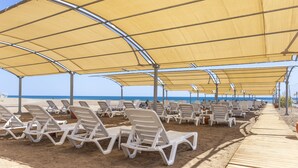 A beachside area with rows of white lounge chairs under large yellow canopies, a wooden walkway, and the ocean in the background. - Palmora Lara Hotel - All inclusive (Antalya)