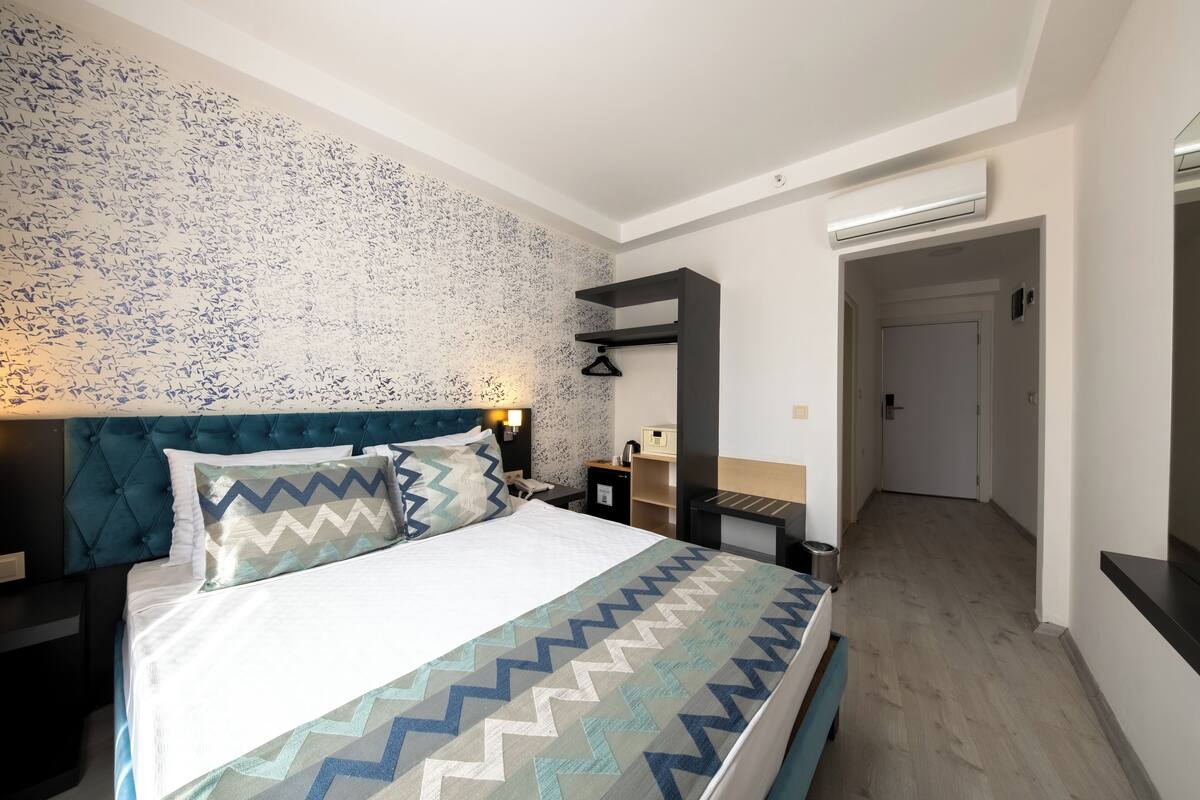 A modern hotel room with a large bed, a headboard, a nightstand, a bedside table, and a wall-mounted air conditioner.
