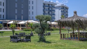 A hotel courtyard with outdoor seating, a thatched gazebo, and modern buildings in the background. - Palmora Lara Hotel - All inclusive (Antalya)