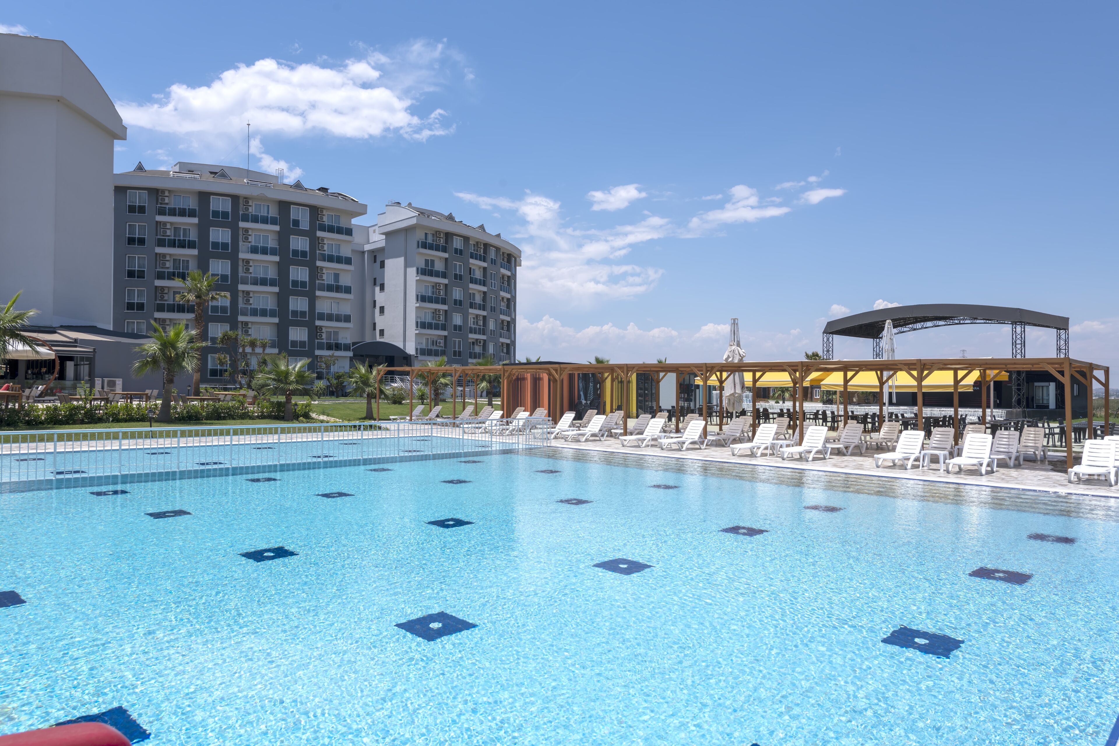 Una piscina con sillas de descanso, una zona de estar cubierta y un edificio de hotel al fondo.