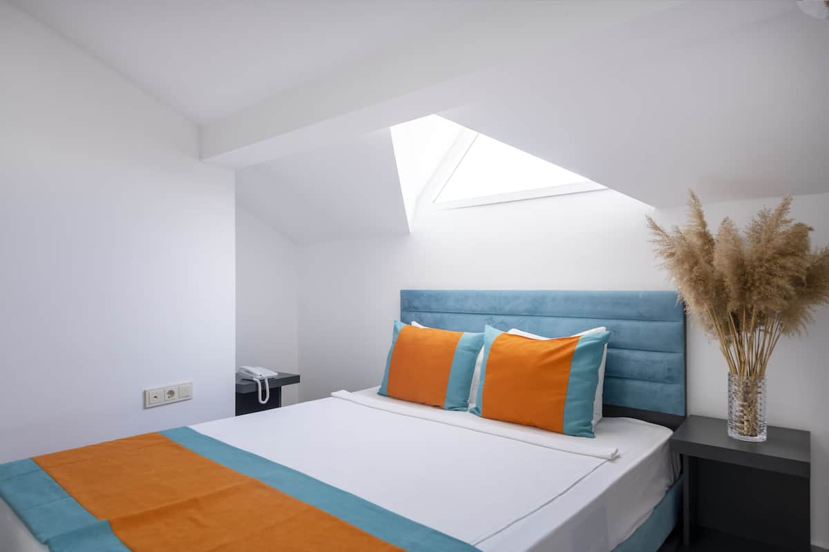 A modern bedroom with a skylight, a bed with orange and blue bedding, and a bedside table with a vase of dried plants.