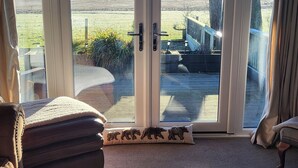 A room with a large sliding glass door leading to an outdoor deck. Inside, there is a brown leather sofa, a small rug, and a table with a vase of flowers. - Cosy Wood Cabin in Rural Area Near National Park (Cumnock)