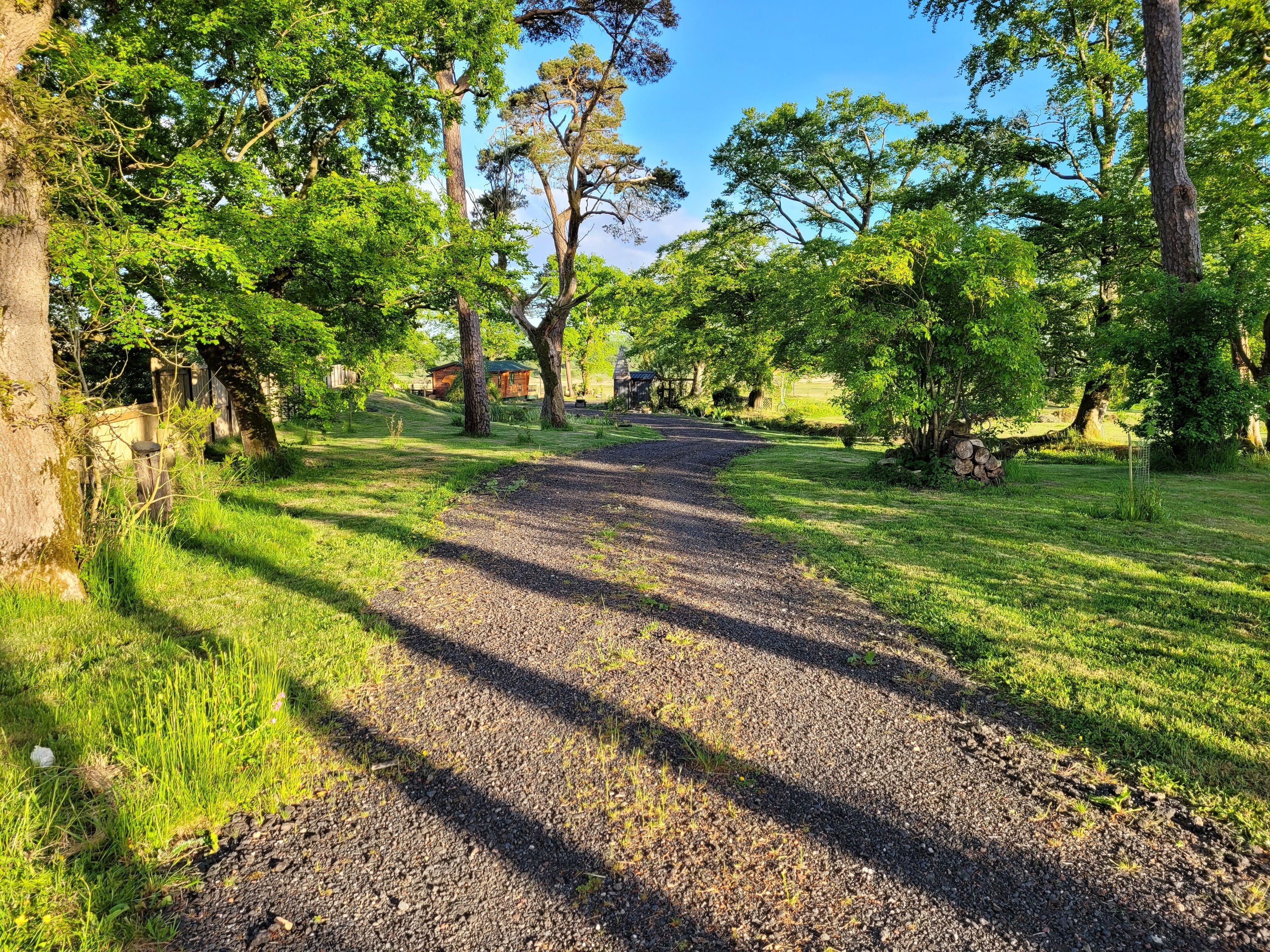 A gravel path leading through a park with trees and a wooden structure in the background.