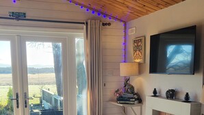 A cozy living room with a fireplace, a television mounted on the wall, and a sliding glass door leading to a scenic outdoor view. - Cosy Wood Cabin in Rural Area Near National Park (Cumnock)
